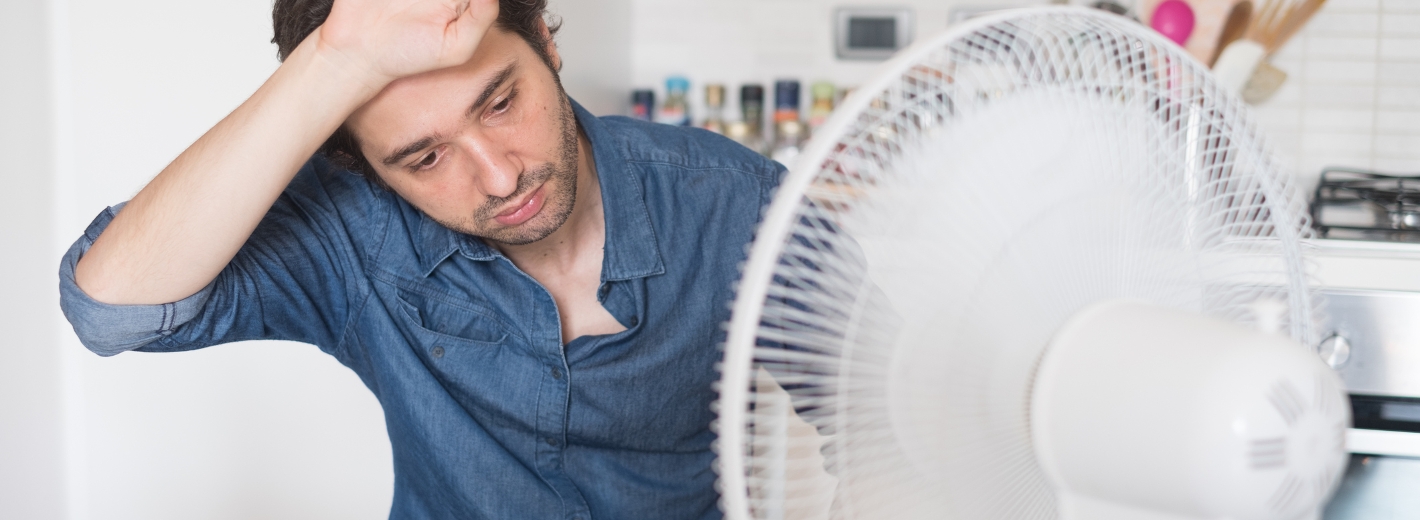Mann wischt sich den Schweiß von der Stirn und sitzt vor einem Ventilator im Büro.