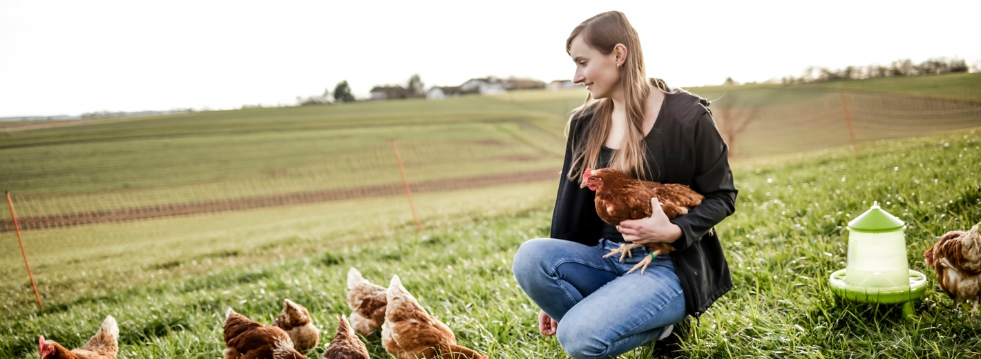 Die Frau kniet auf einer Wiese mit Hühnern und hält ein Huhn in der Hand