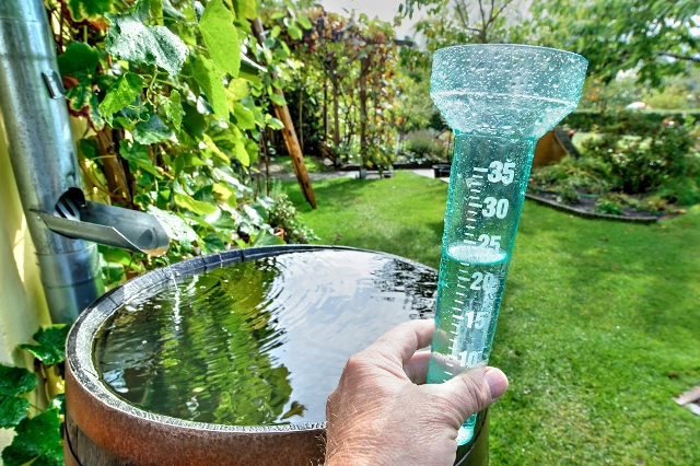 Ein Regenmesser, vor einer Regentonne mit einem Fallrohr, zeigt die Niederschlagsmenge an